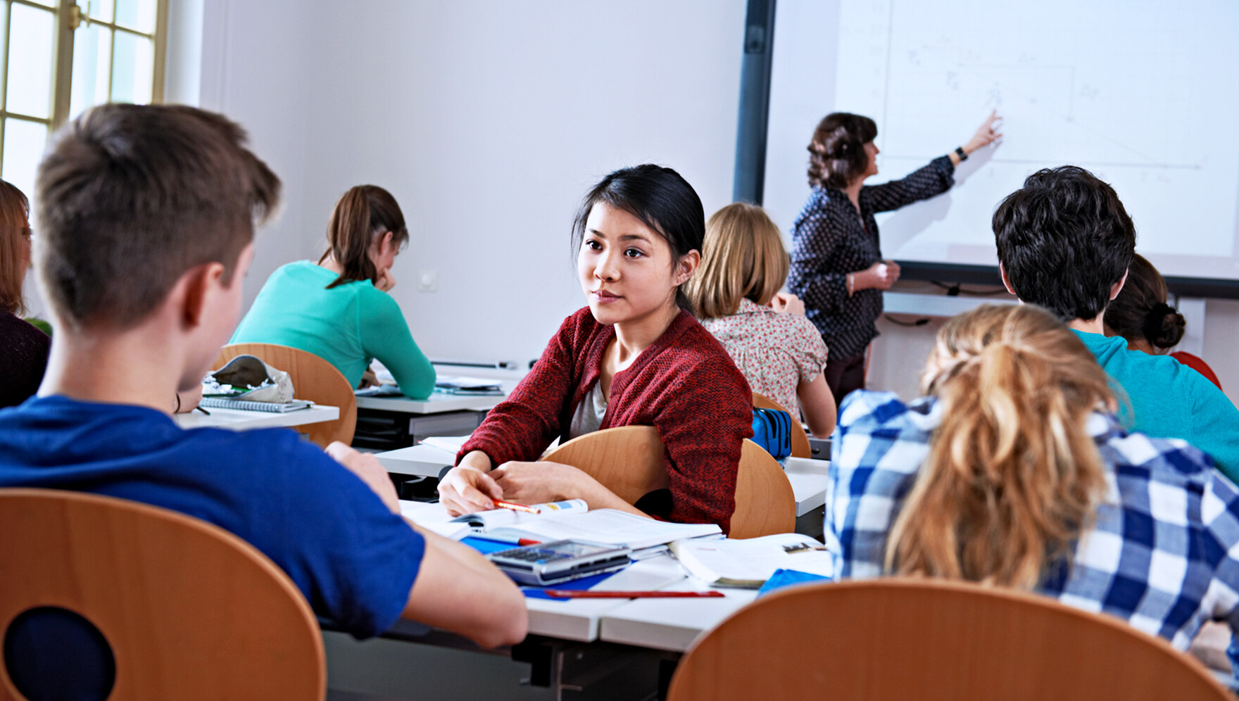 Szene aus einem Klassenzimmer: Die Lehrerin zeigt etwas am Projektor, ein Mädchen dreht sich zu ihren Klassenkameraden nach hinten um.